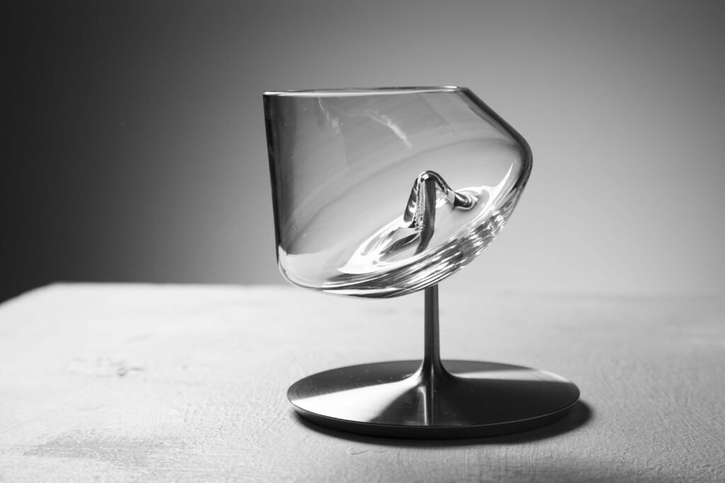 Fotografía en blanco y negro de la copa cónica inclinada sobre su pie metálico, mostrando el pico central en el fondo del cristal que le permite mantener el equilibrio pese a su forma asimétrica.