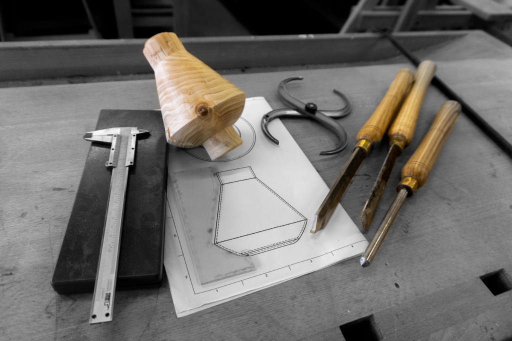 Fotografía en blanco y negro de un boceto en papel con la plantilla del perfil de la copa, junto a un modelo de madera torneado, un calibre y gubias de torno sobre una mesa de taller.
