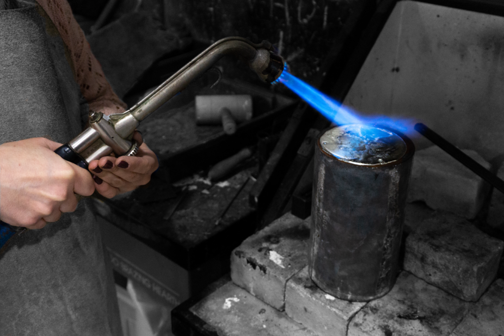 Fotografía en blanco y negro de una persona dirigiendo la llama azul de un soplete hacia un cilindro metálico apoyado sobre bloques refractarios, en un proceso de fundición o soldadura.