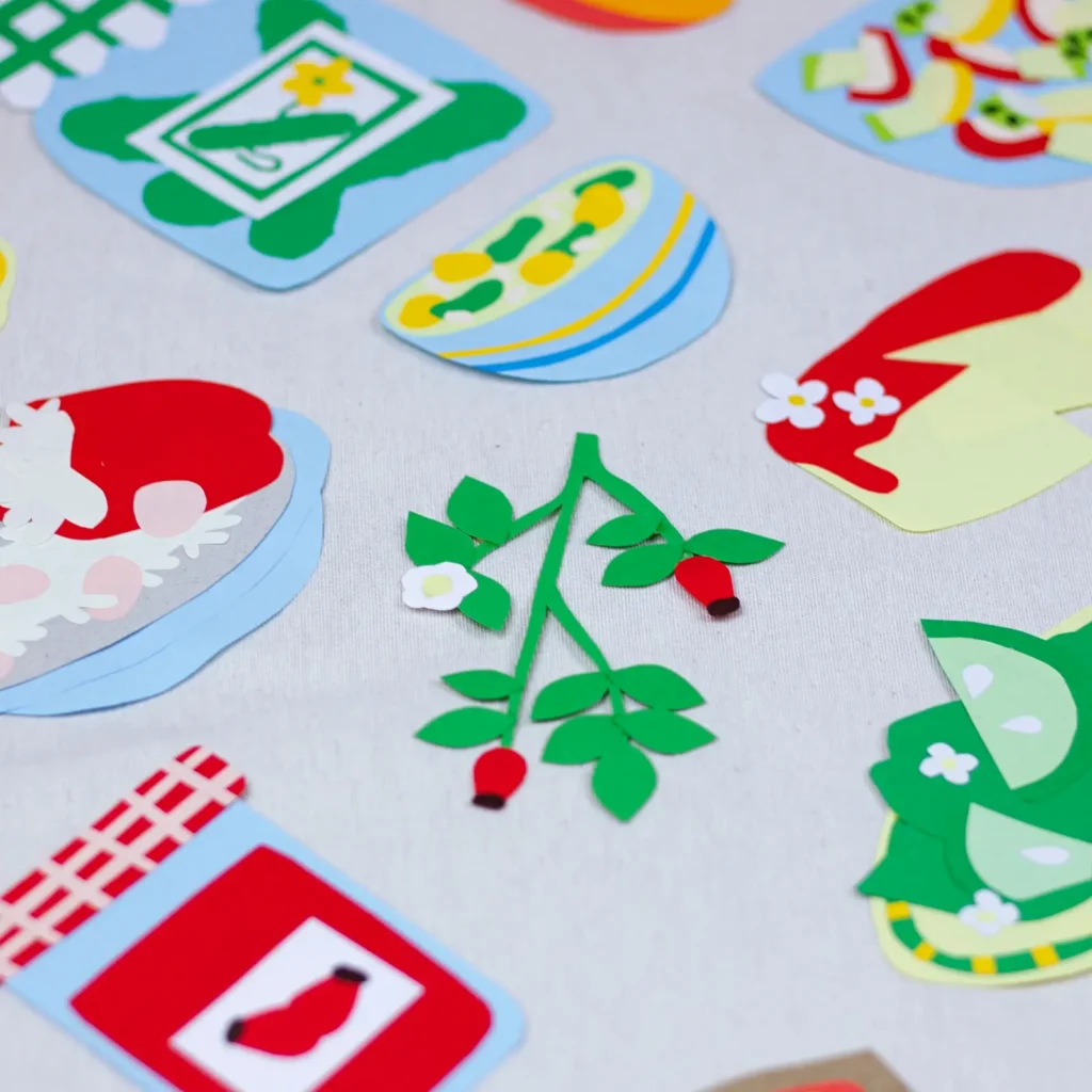 Detalle de las figuras de papel recortadas que decoran el fondo de tela: platos de comida, tarros de conserva y una ramita con flores y frutos rojos.