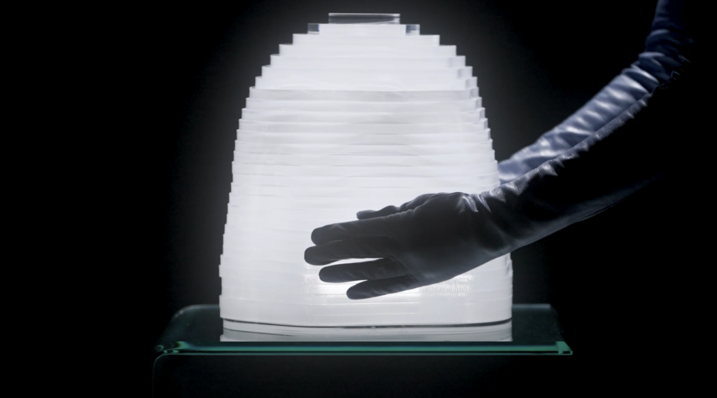 Una mano enguantada de negro toca la superficie estriada de la lámpara "cápsula del tiempo", iluminada desde dentro con luz blanca, sobre un fondo completamente oscuro.