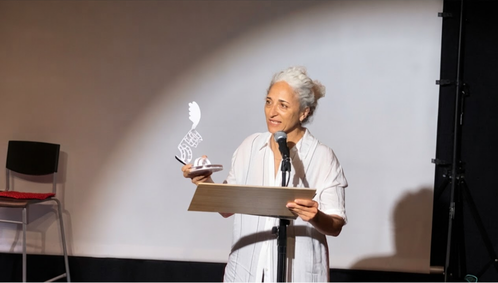 Una mujer con el pelo canoso, vestida de blanco, sostiene uno de los trofeos con forma de ala mientras habla ante un atril, con una silla vacía y una pantalla de fondo en la sala del festival.