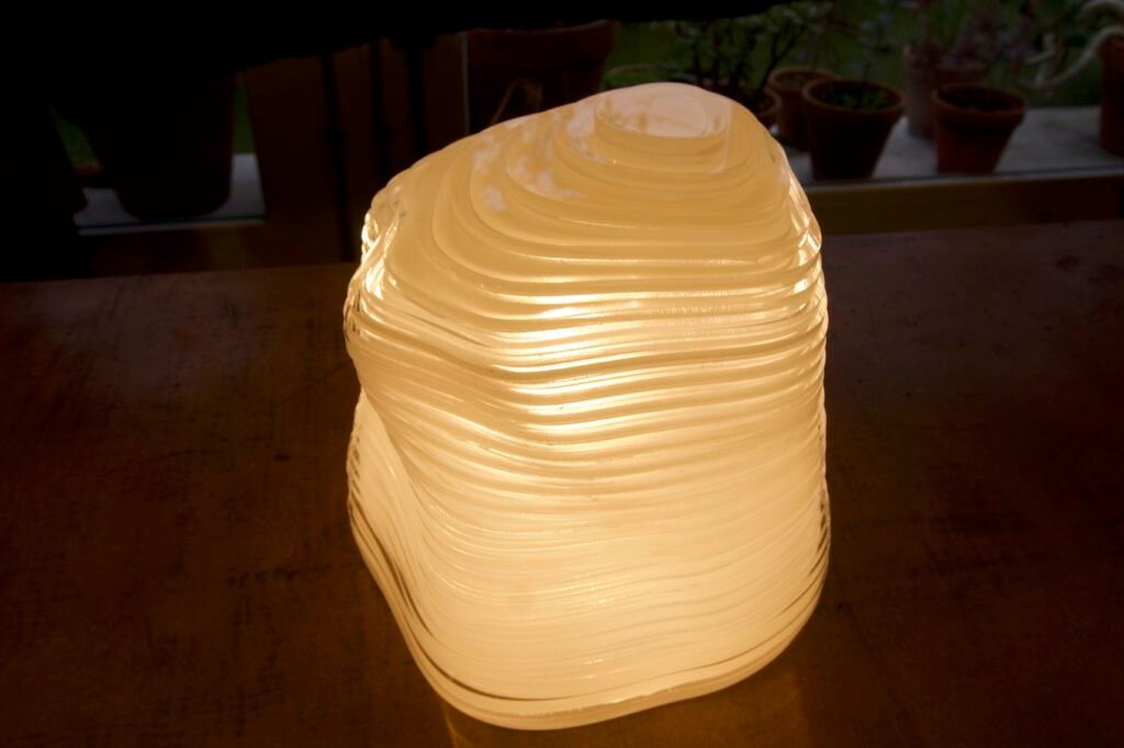 La lámpara "cápsula del tiempo" encendida sobre una mesa de madera junto a una ventana con macetas, mostrando su textura de capas onduladas y translúcidas iluminadas desde dentro.