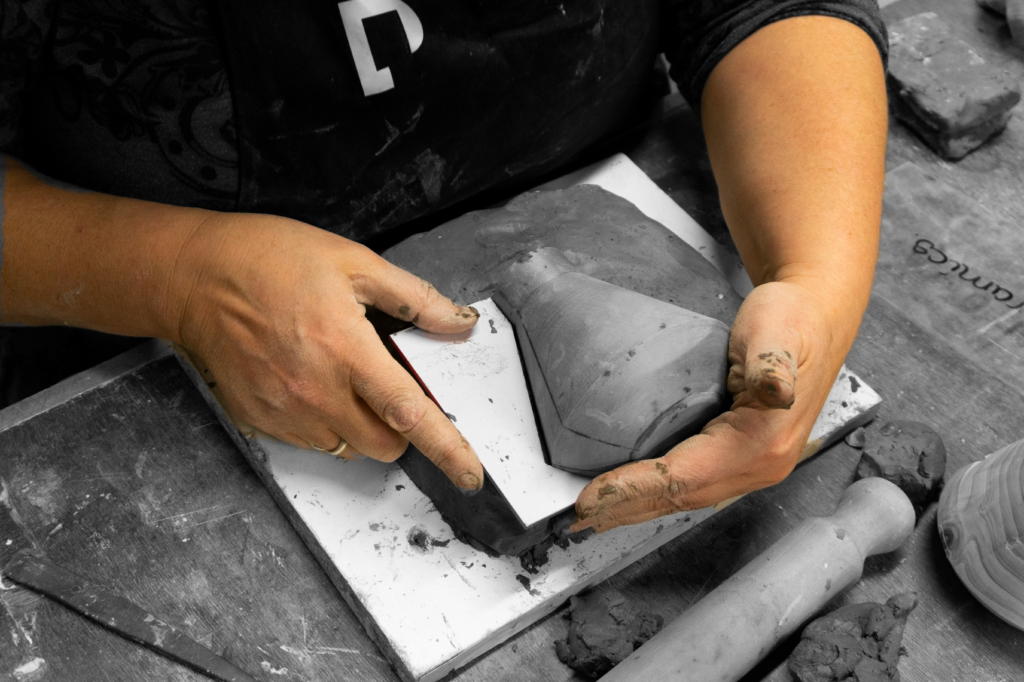 Fotografía en blanco y negro de dos manos moldeando una pieza de arcilla gris con forma cónica sobre una plantilla de cartón, con herramientas de modelado alrededor.