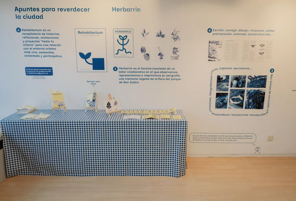 Panel expositivo titulado "Apuntes para reverdecer la ciudad — Herbarrio", con las cubiertas de ambas publicaciones, ilustraciones botánicas y fotografías del proceso, sobre una mesa con mantel de cuadros azules donde se exponen ejemplares y tarjetas.