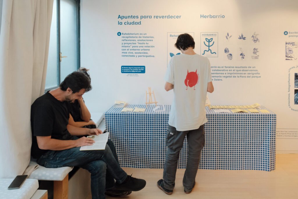 Dos personas consultan el material expositivo del proyecto: una sentada leyendo un ejemplar del fanzine, y otra de pie mirando los documentos sobre la mesa con mantel de cuadros, frente al panel "Apuntes para reverdecer la ciudad".