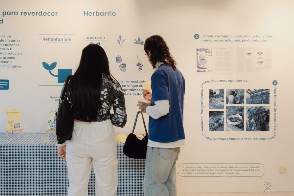 Dos personas de espaldas observan el panel expositivo "Apuntes para reverdecer la ciudad — Herbarrio" con sus ilustraciones botánicas y fotografías del proceso.