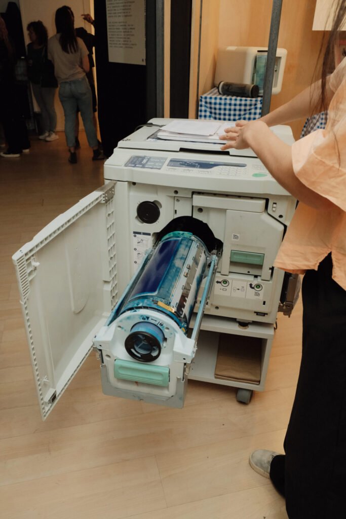 Detalle de la máquina risográfica con el tambor de tinta azul abierto y visible, mientras una persona manipula unos papeles sobre la tapa del equipo.