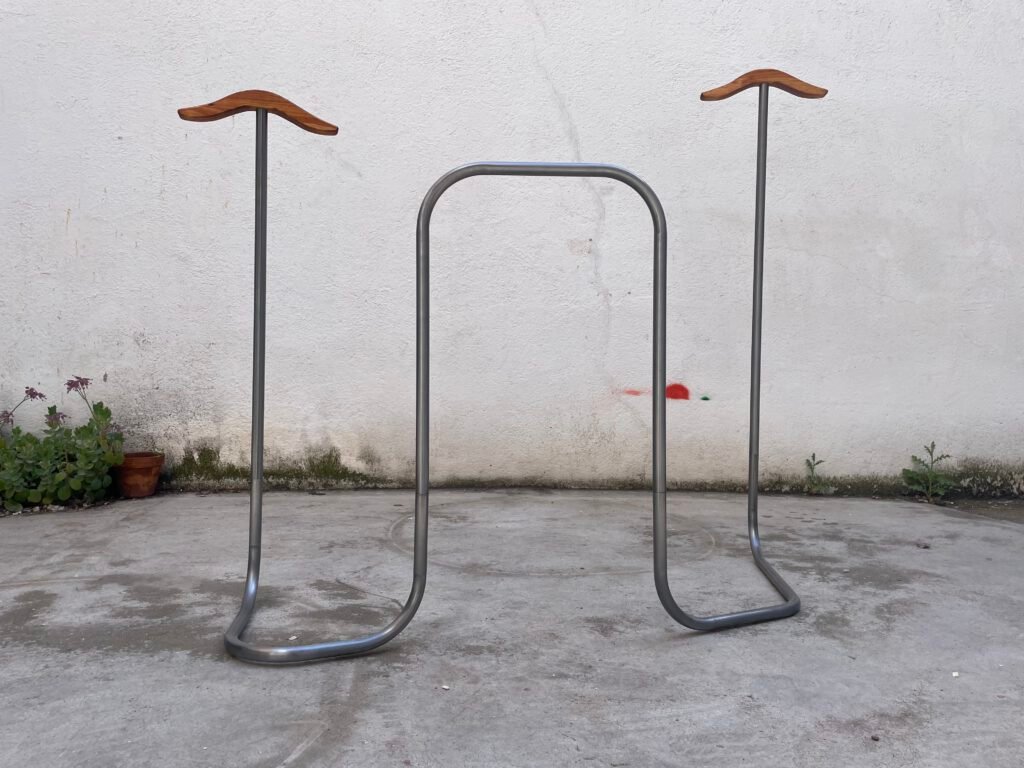 Dos percheros metálicos independientes con reposahombros de madera curvada, unidos por una barra metálica horizontal, ante una pared blanca con macetas y plantas al fondo.