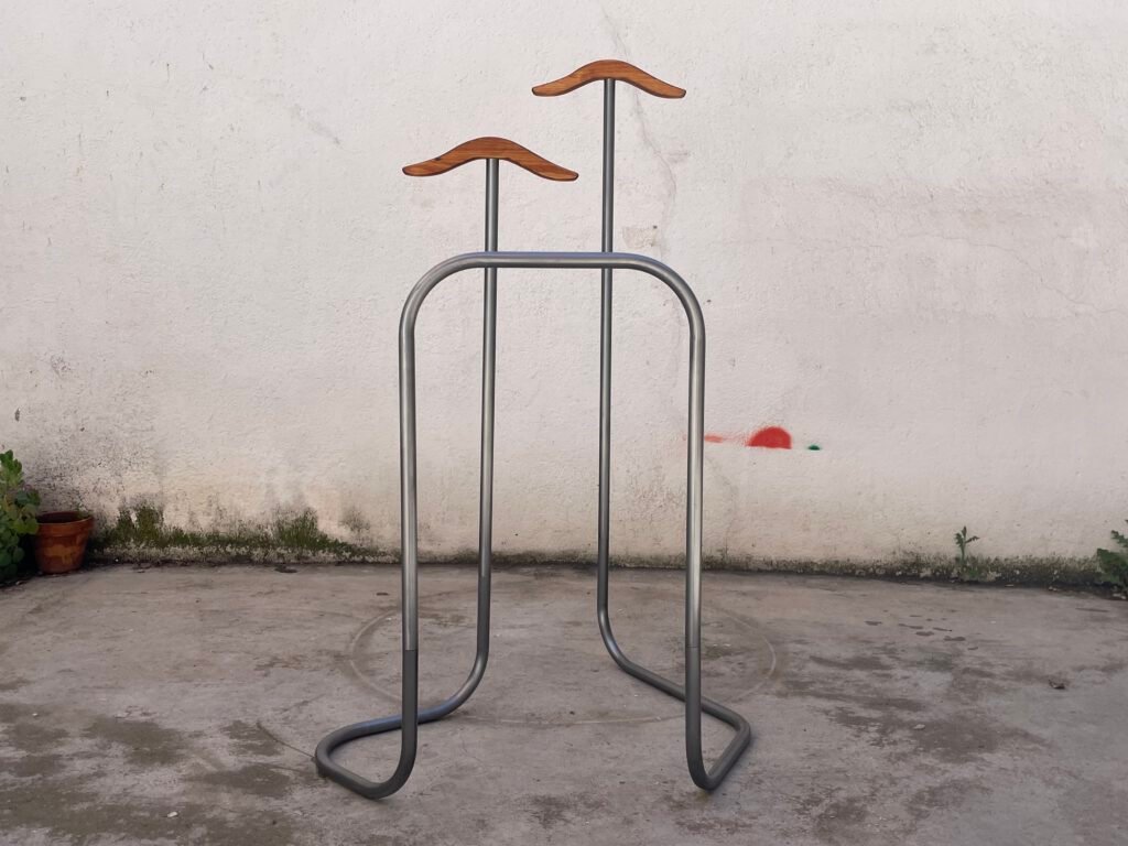Un perchero expositor de estructura metálica en forma de U invertida con dos reposahombros de madera curvada a distinta altura, sobre una pared blanca desgastada.