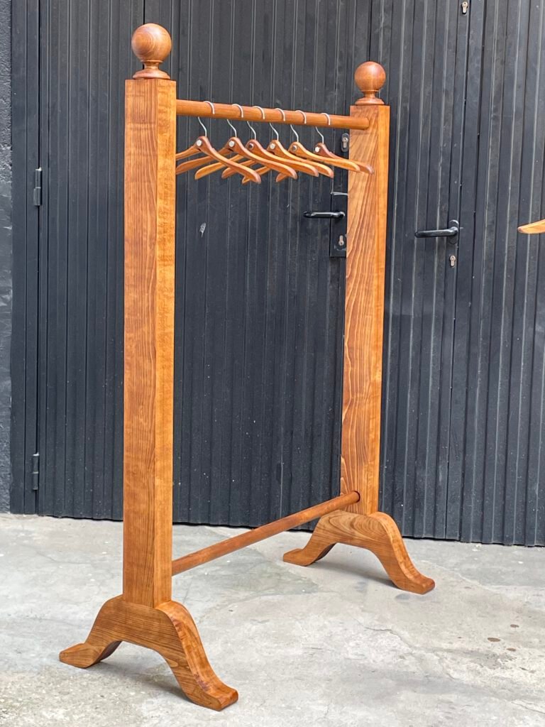 Perchero de madera clara con seis perchas de madera colgadas de una barra horizontal entre dos remates esféricos, apoyado sobre una base con patas curvas, ante una puerta metálica oscura.