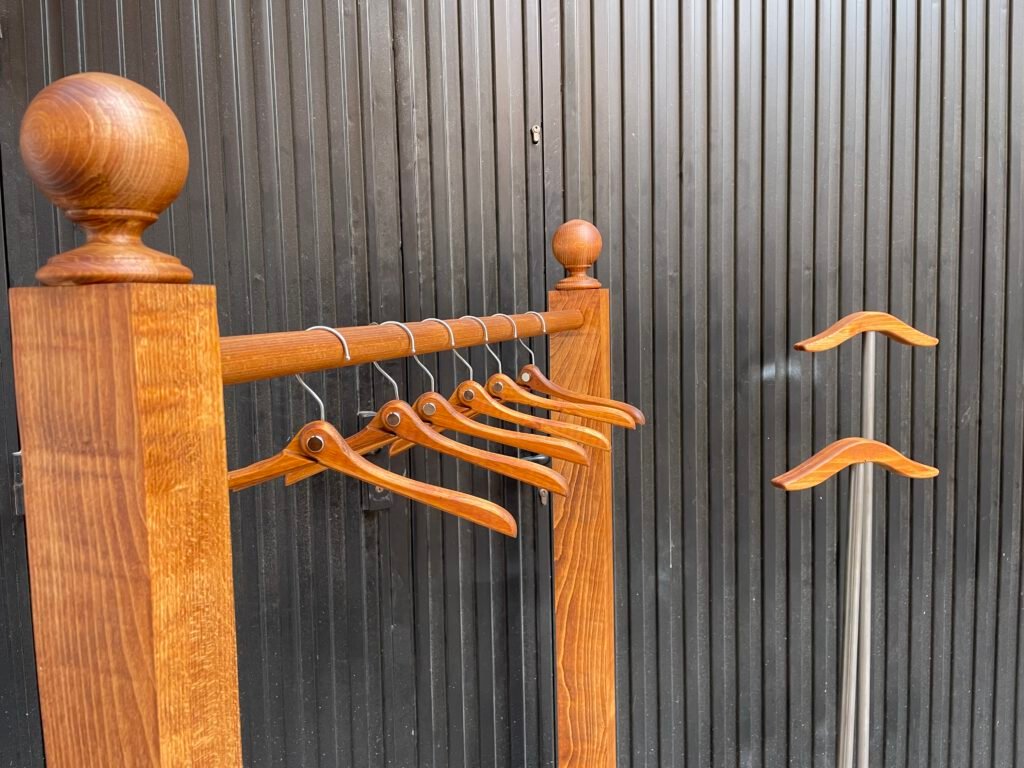 Detalle del remate esférico de madera y la barra con perchas de madera colgadas del perchero, con otro perchero metálico de reposahombros al fondo.