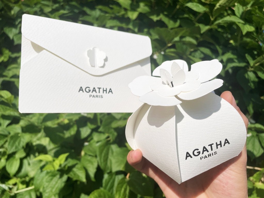 Sobre y caja de regalo de papel blanco con el logo de Agatha Paris y un cierre troquelado en forma de flor; una mano sostiene la caja, rematada con una flor de papel, sobre un fondo de hojas verdes.