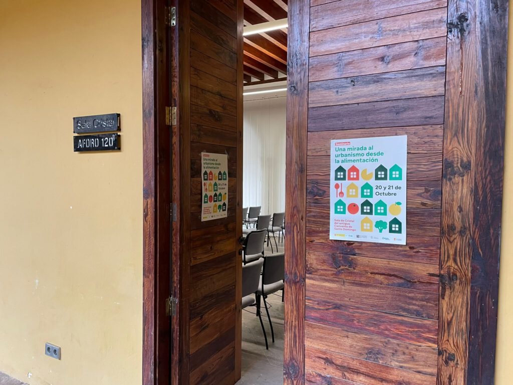 Puerta de madera de la Sala Cristal (aforo 120) con el cartel del seminario impreso y pegado, y sillas colocadas dentro de la sala al fondo.