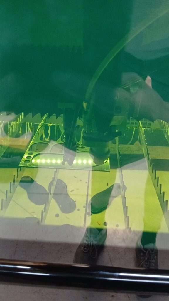 Vista a través de un panel de metacrilato verde de varias piezas del trofeo recién cortadas con láser, con los pies de quien fotografía reflejados en la superficie.