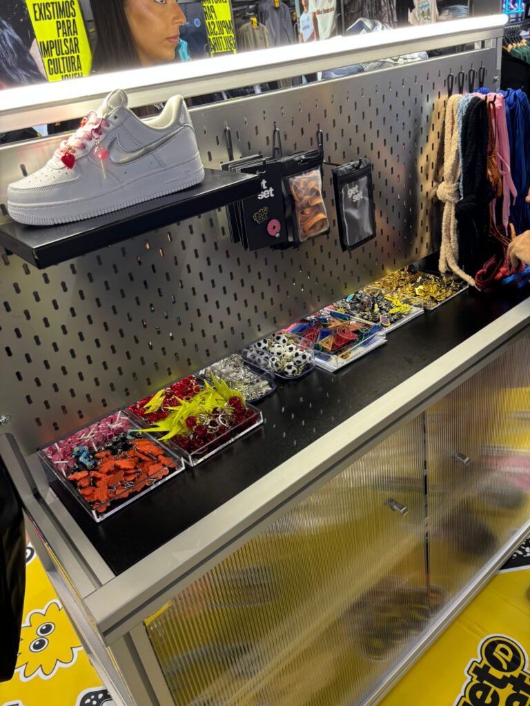 Bandejas de abalorios y colgantes de colores para personalizar zapatillas, dispuestas sobre el mostrador del mueble "get set", con una Nike Air Force 1 blanca decorada con charms en la balda superior.