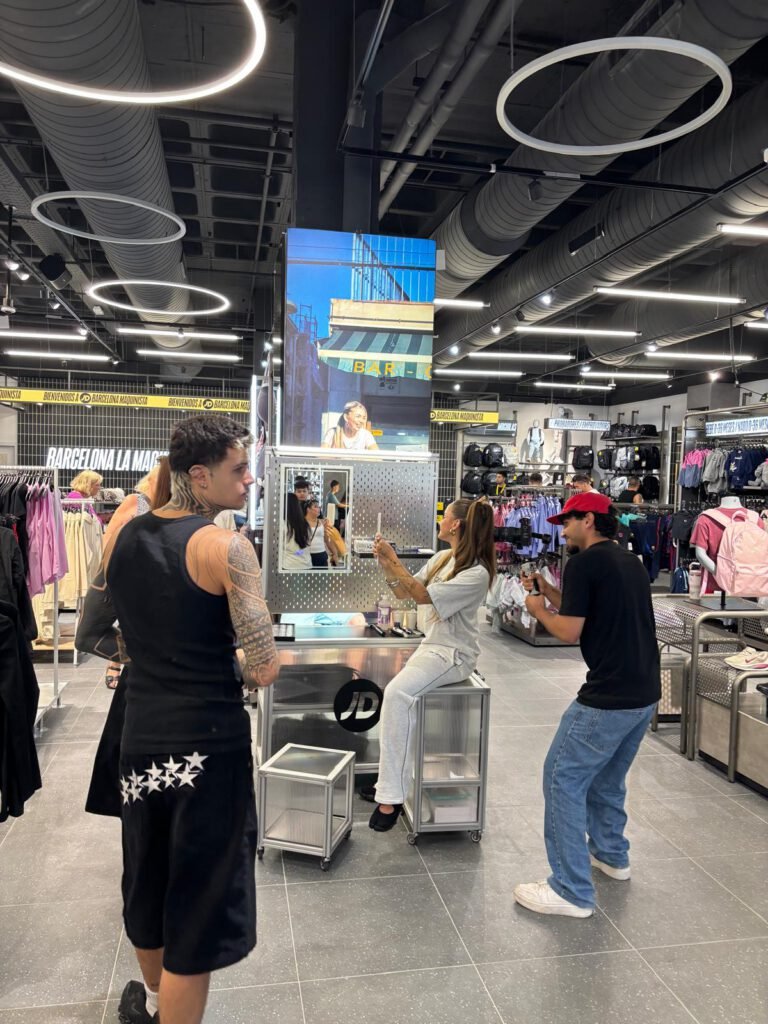 Vista general de la tienda JD Sports con gente reunida alrededor del mueble "get set", una pantalla con vídeo sobre el expositor, y una persona sentada en él siendo grabada por un compañero.