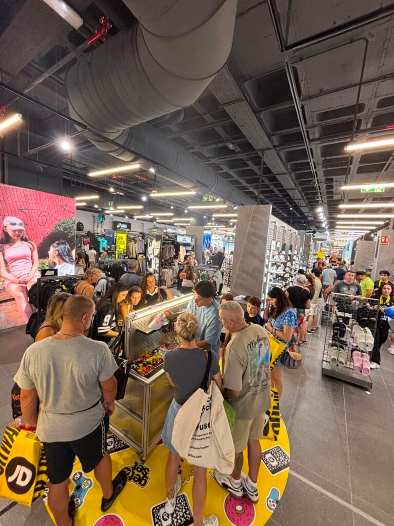 Grupo numeroso de clientes alrededor del mueble "get set", colocado sobre una plataforma circular amarilla con pegatinas decorativas, en medio de la tienda llena de gente.