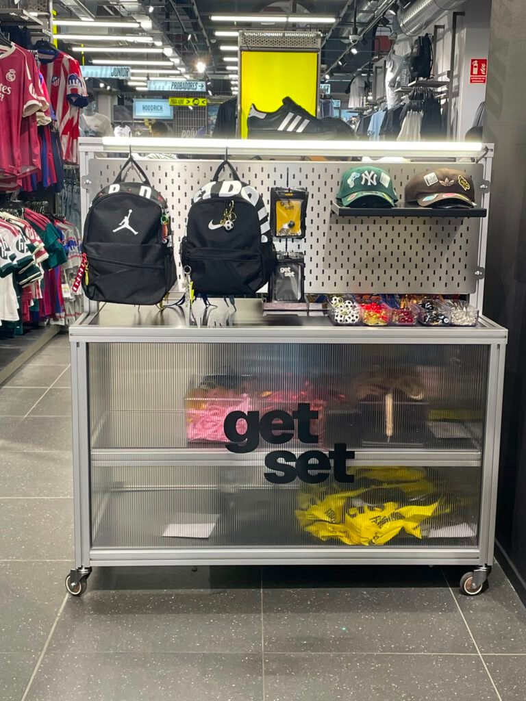 Mueble expositor metálico con ruedas y el rótulo "get set" en el frontal traslúcido, con mochilas Jordan y Nike, gorras y complementos colgados en un panel perforado, dentro de una tienda de deporte.