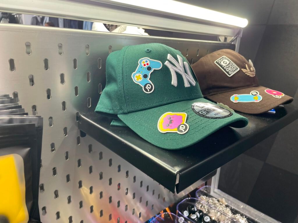 Dos gorras personalizadas con chapas decorativas (un mando de videoconsola, un monopatín, un personaje) sobre una balda del expositor: una verde con el logo de los Yankees de Nueva York y otra marrón de Adidas.