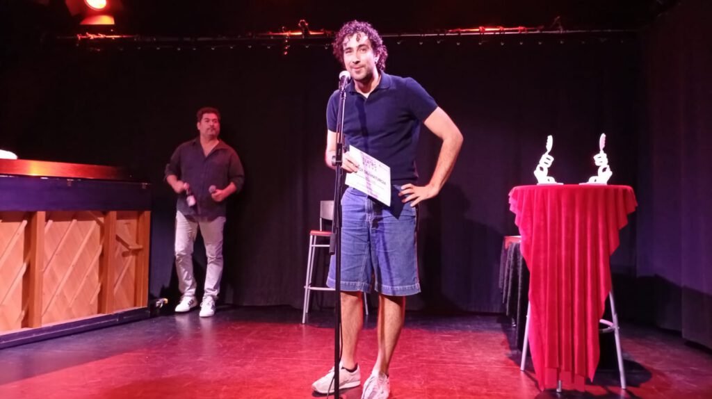 Un hombre con pantalón corto vaquero habla ante un micrófono en el escenario de una sala de teatro, con dos trofeos blancos sobre una mesa cubierta de tela roja al fondo.