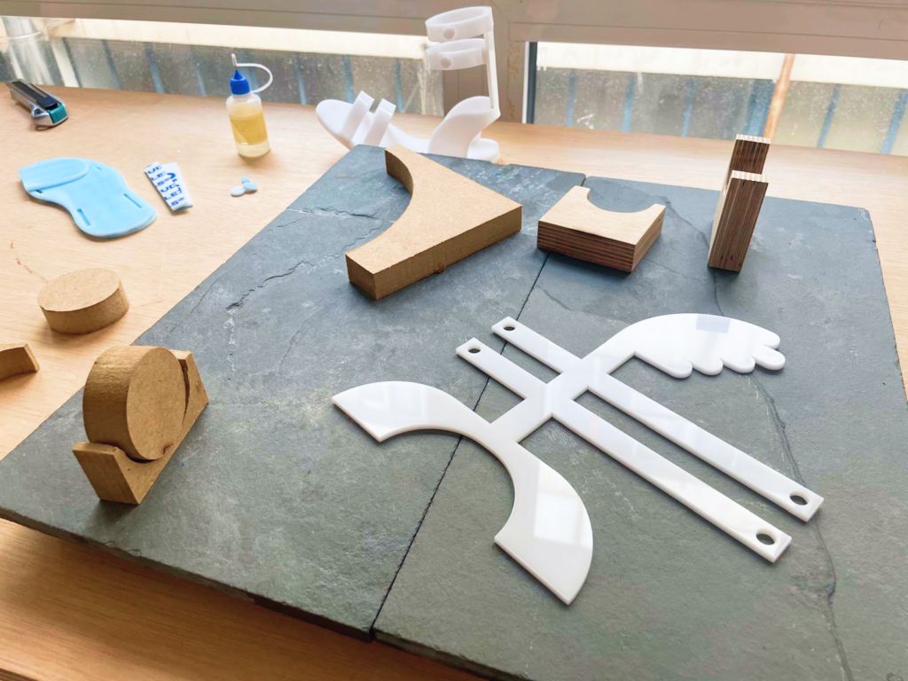 Mesa de taller con piezas cortadas de madera y plástico blanco en forma de ala, moldes, pegamento y herramientas, junto a un boceto del trofeo apoyado al fondo.