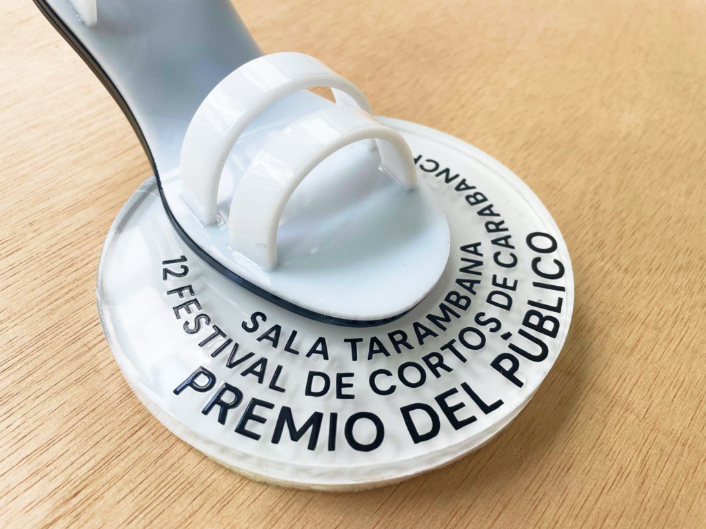 Detalle de la peana circular transparente de uno de los trofeos, con el texto grabado "12 Festival de Cortos de Carabanchel — Premio del Público" y la base blanca de la sandalia apoyada encima.