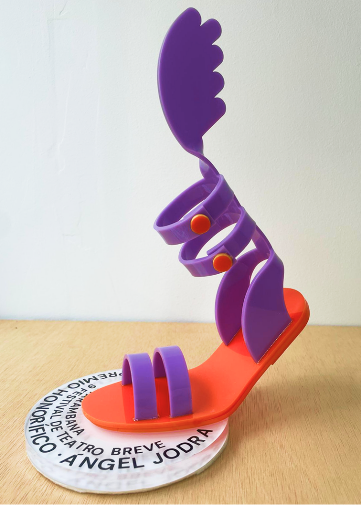 Detalle del trofeo en morado y naranja con forma de sandalia alada, con una correa de tobillo enrollada y remaches de color naranja, sobre una peana circular con el texto "Premio Honorífico · Ángel Jodra".