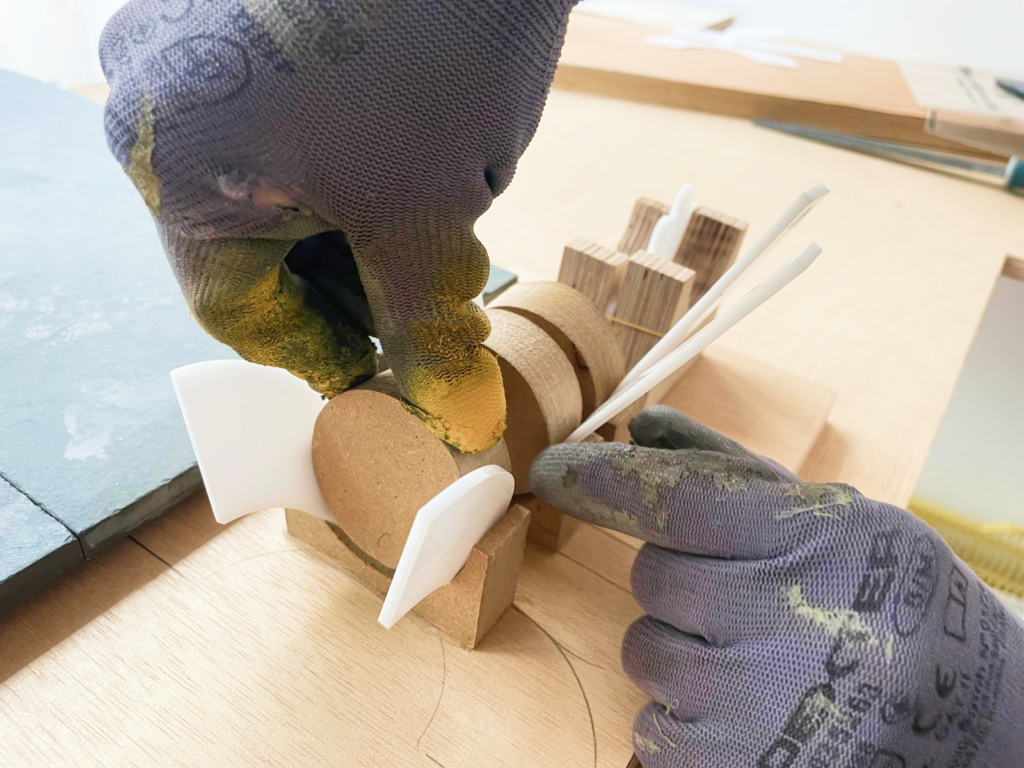 Unas manos con guantes de trabajo ensamblan piezas de madera y plástico blanco curvado sobre una mesa de taller, en el proceso de montaje del trofeo.