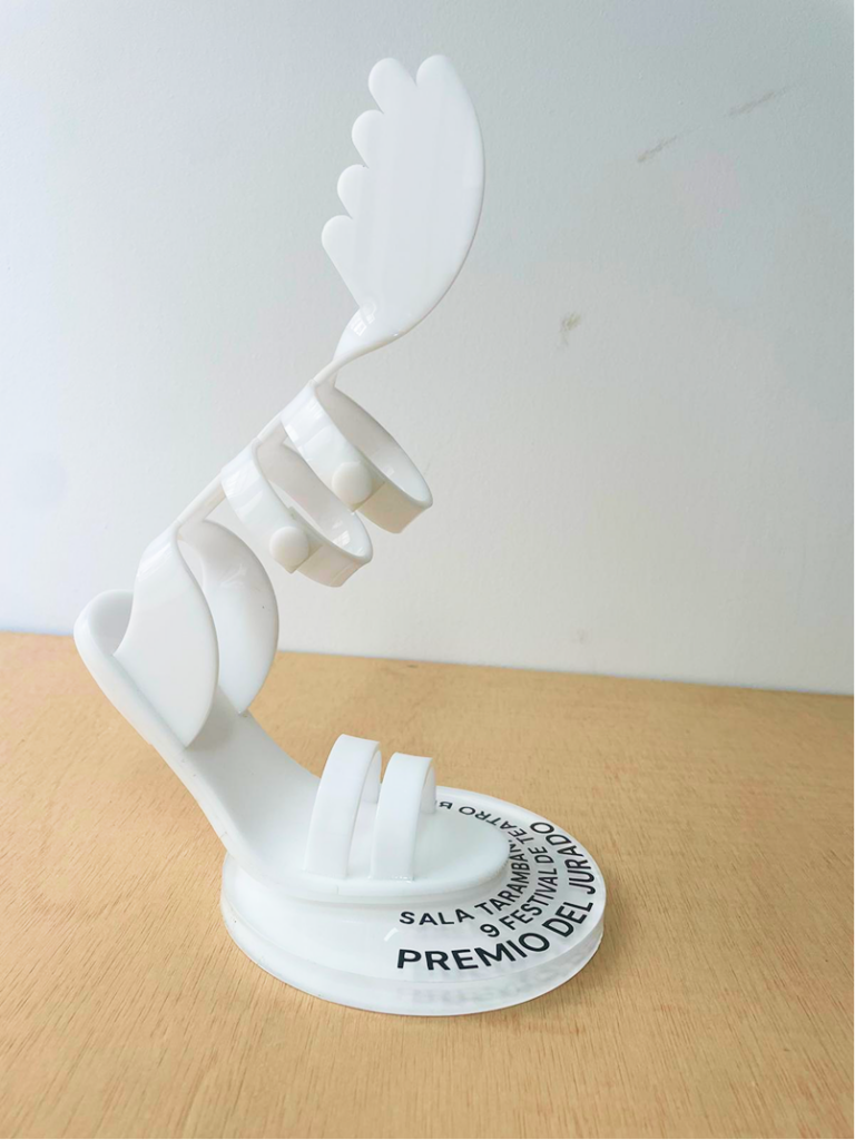 El trofeo en blanco con forma de sandalia alada, con la correa de tobillo enrollada en espiral, sobre una peana circular con el texto "Sala Tarambana — Premio del Jurado".