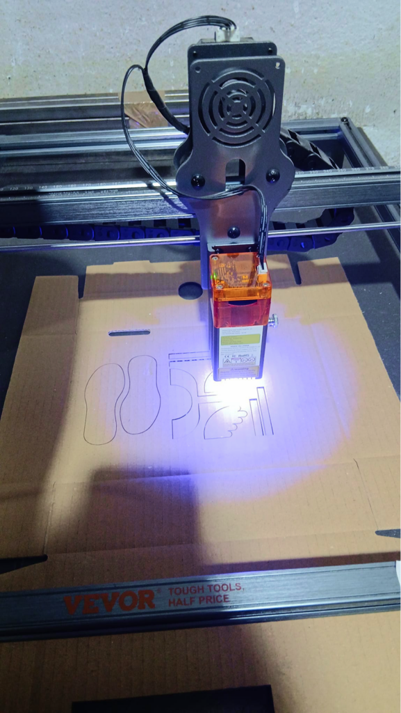 Una máquina de corte láser grabando el contorno de una pieza en forma de ala sobre una plancha de cartón, con el haz violeta iluminando la superficie.