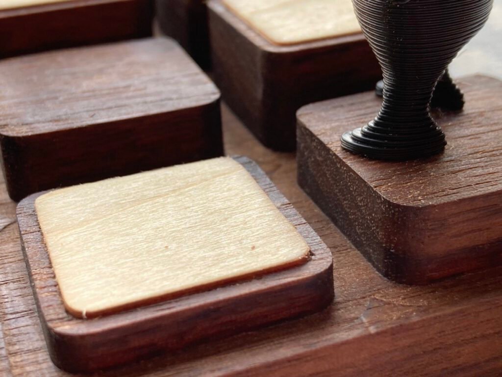 Detalle en primer plano de las casillas de madera del tablero, con las vetas de la madera clara a la vista, y la base de una pieza negra impresa en 3D apoyada sobre una de ellas.