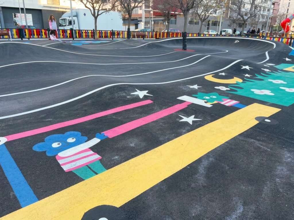 Tramo ondulado del circuito con ilustraciones de personajes de colores jugando con cintas rosas y amarillas entre estrellas y una luna, junto a una zona verde con nubes y motivos florales.