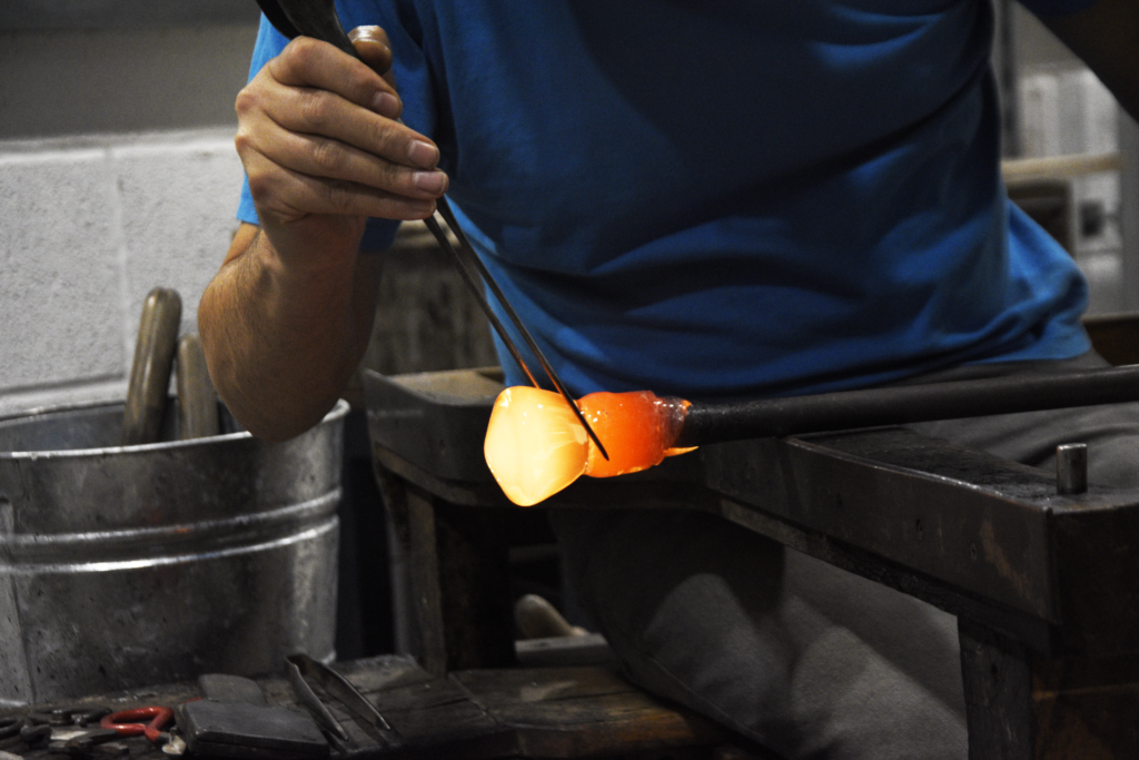 Un artesano sostiene con unas pinzas una pieza de vidrio incandescente en naranja y amarillo sobre la caña del soplado, junto a un cubo metálico con herramientas.