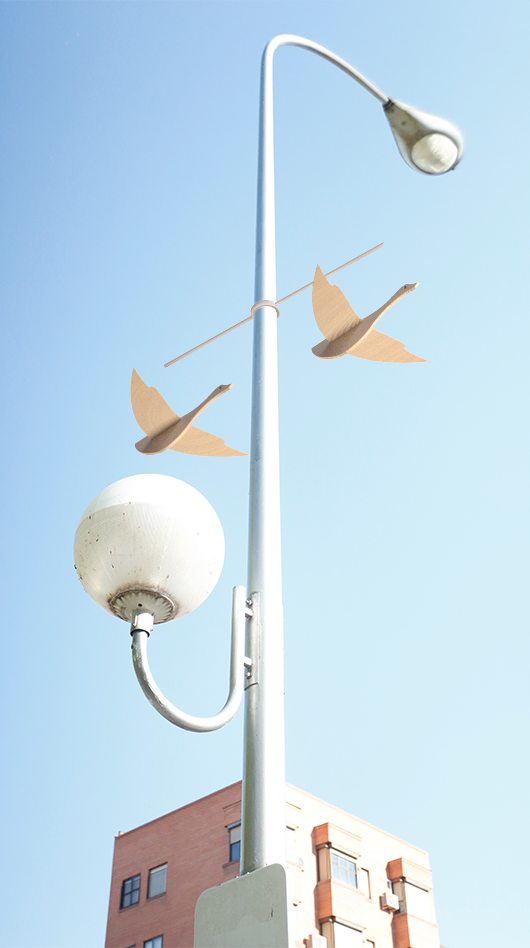 Render de una farola urbana con dos siluetas de aves de madera clara colgadas de una varilla horizontal, simulando el vuelo, junto a un edificio de fondo bajo un cielo azul.