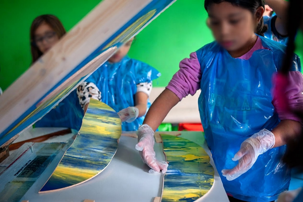 Dos niñas con delantales azules de plástico manipulan piezas de madera pintadas con manchas de pintura azul y amarilla, apoyadas en una mesa con fondo verde, durante un taller.
